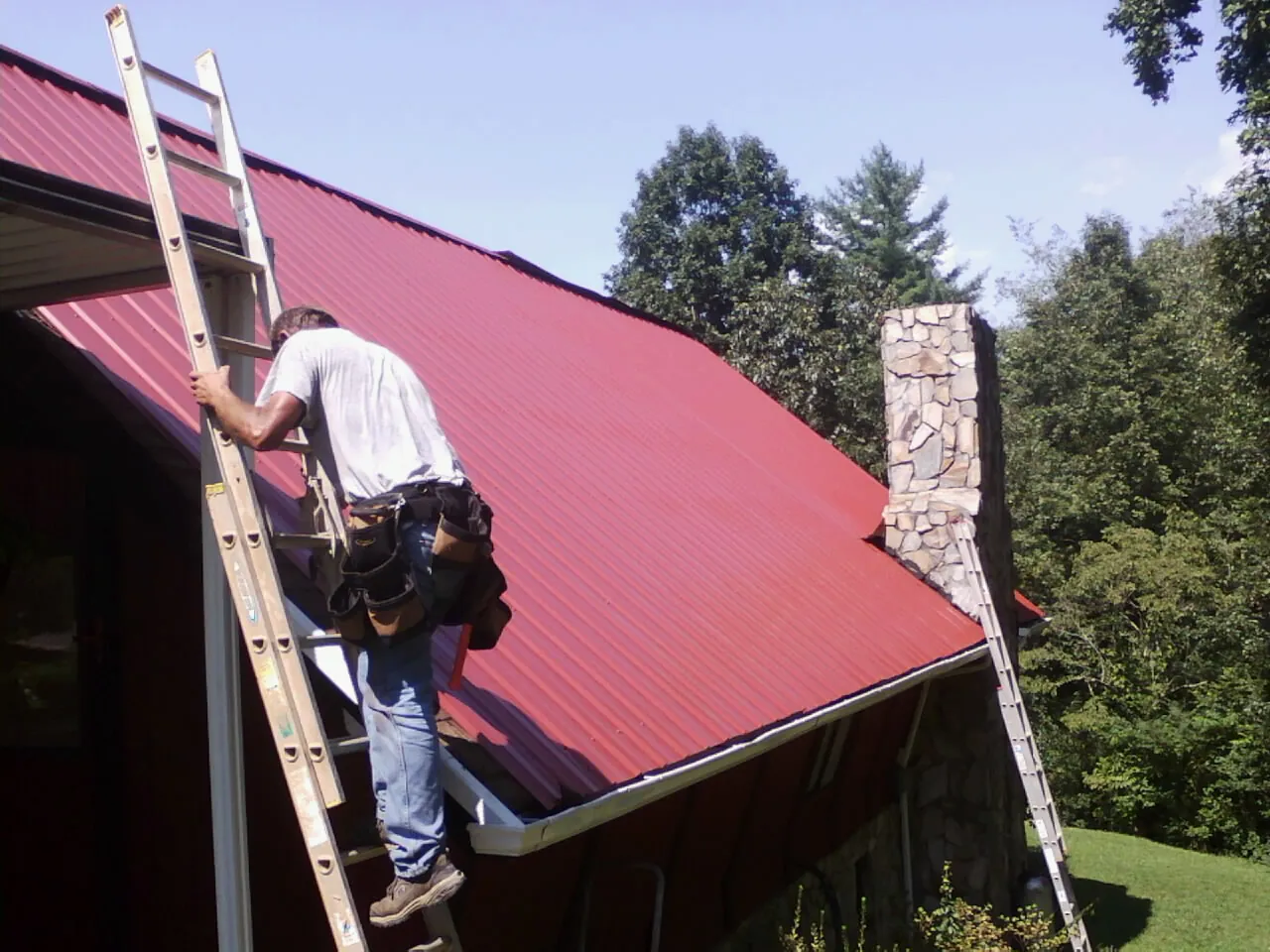 Roofer on a ladder beside a red metal roof and stone chimney — Soffit & Fascia Repair by Roofing Brothers in Etoile